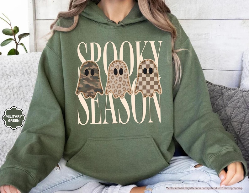 May include: A military green hooded sweatshirt featuring the text SPOOKY SEASON in large cream letters. Centered between the words are three ghost illustrations with black oval eyes, each featuring a different pattern: camouflage, leopard print, and a brown checkerboard.