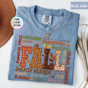 以下が含まれることがあります： 様々な模様と質感で「FALL」の文字がプリントされた水色のTシャツ。 「Pumpkin Spice」、「Sweater Weather」、「Giving Thanks」など、秋をテーマにした言葉がプリントされています。 金色のネックレスがTシャツの上に置かれています。