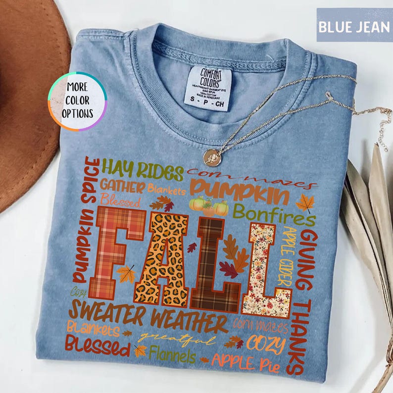 以下が含まれることがあります： 様々な模様と質感で「FALL」の文字がプリントされた水色のTシャツ。 「Pumpkin Spice」、「Sweater Weather」、「Giving Thanks」など、秋をテーマにした言葉がプリントされています。 金色のネックレスがTシャツの上に置かれています。