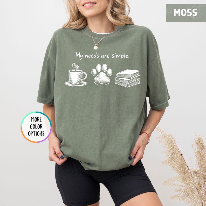 May include: Moss green t-shirt with the phrase "My needs are simple." printed in white. The graphic includes a coffee cup, a paw print, and a stack of books. The shirt is a relaxed fit.