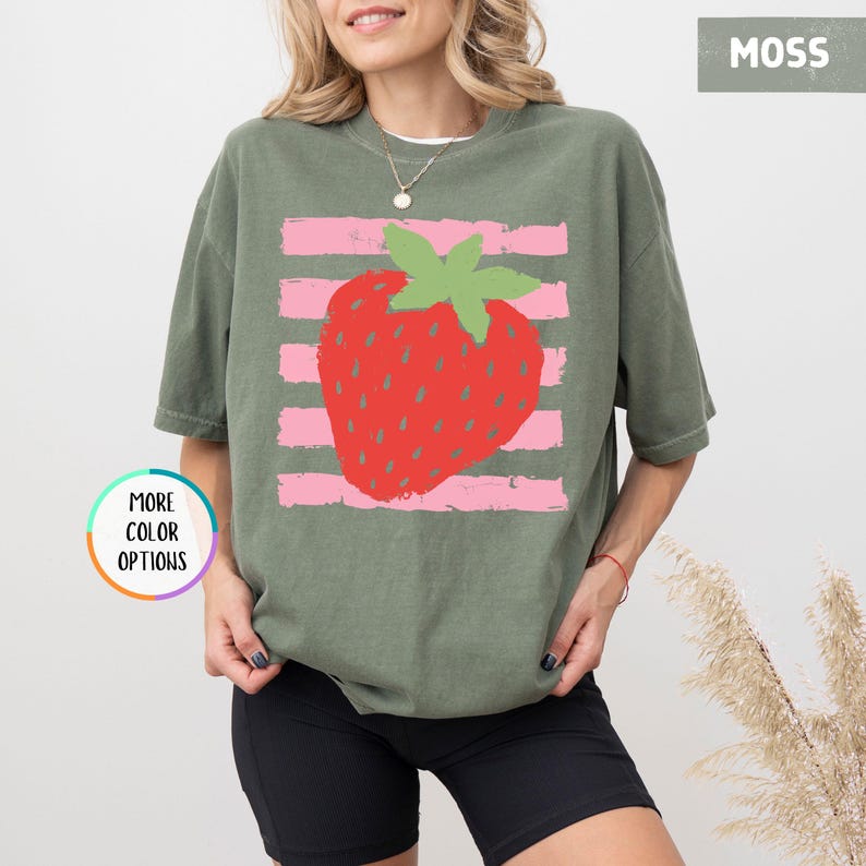 May include: A moss green short-sleeve t-shirt featuring a large red strawberry graphic with a green stem centered over horizontal light pink stripes. The shirt has a crew neckline and a relaxed fit. A small circular badge reads MORE COLOR OPTIONS.