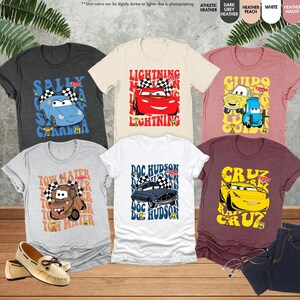以下が含まれることがあります： 漫画の車のデザインが施されたTシャツのコレクション。Tシャツは、グレー、白、バーガンディなど、さまざまな色があります。各シャツには、「Lightning」や「Tow Mater」などのテキストとともに、異なる車のキャラクターが表示されています。靴とジーンズも写っています。