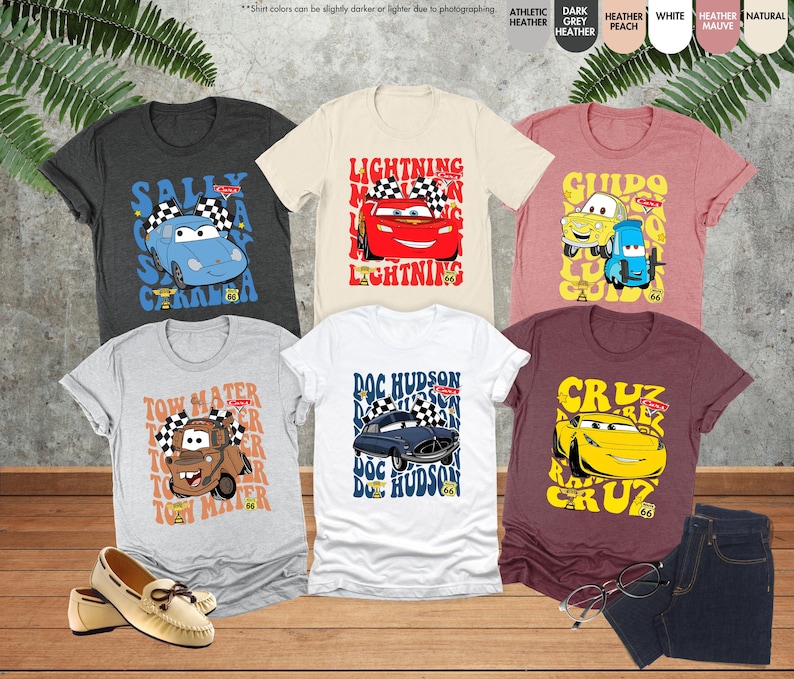 以下が含まれることがあります： 漫画の車のデザインが施されたTシャツのコレクション。Tシャツは、グレー、白、バーガンディなど、さまざまな色があります。各シャツには、「Lightning」や「Tow Mater」などのテキストとともに、異なる車のキャラクターが表示されています。靴とジーンズも写っています。