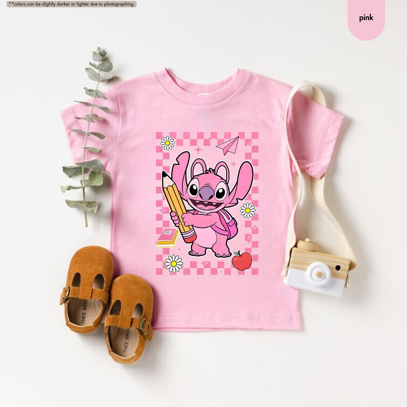 May include: A pink t-shirt with a cartoon character holding a pencil, backpack, and apple design. The design is set against a checkered background with daisies and a paper airplane. The shirt is paired with brown leather shoes.