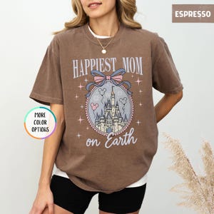 May include: Espresso-colored t-shirt with the text "Happiest Mom on Earth" above a castle graphic. The graphic includes a pink bow, Mickey Mouse balloons, and stars. The shirt has short sleeves and a crew neck.