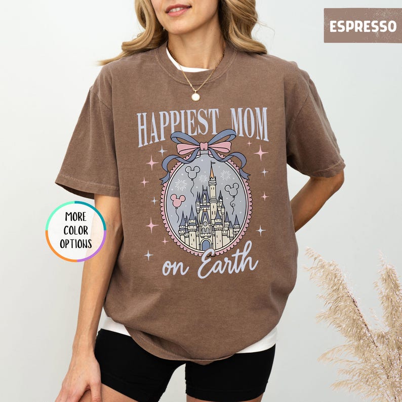 May include: Espresso-colored t-shirt with the text "Happiest Mom on Earth" above a castle graphic. The graphic includes a pink bow, Mickey Mouse balloons, and stars. The shirt has short sleeves and a crew neck.