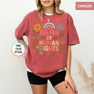 May include: Crimson-colored t-shirt with the text "BIG FAN OF HUMAN RIGHTS" in colorful letters. The design includes flowers and a rainbow. The shirt is a comfortable, casual style.