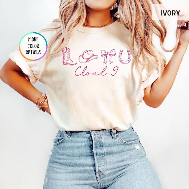 May include: Ivory-colored t-shirt with a pink graphic of a cowboy boot, hat, bow, and horseshoe, with the text "Cloud 9." The shirt has short sleeves and a crew neck. The words "More Color Options" are in a circle on the left.