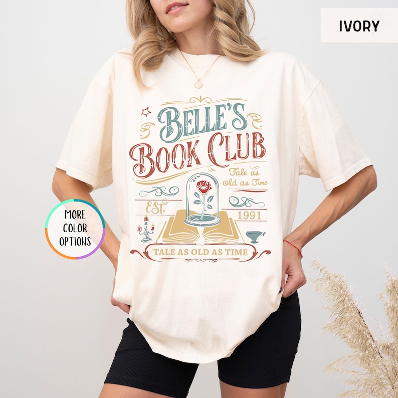 Pu&ograve; includere: T-shirt color avorio con la scritta "Belle's Book Club" in un design vintage. La grafica include una rosa in una cupola di vetro, un libro e la frase "Tale as old as Time". La maglietta presenta anche l'anno "1991" e la parola "EST."