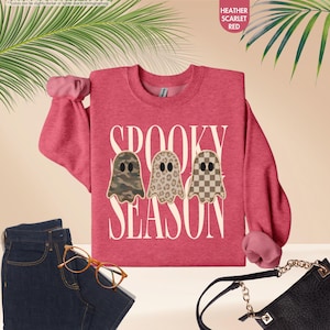 May include: A heather scarlet red crewneck sweatshirt featuring the white text SPOOKY SEASON and three ghost illustrations with camouflage, leopard print, and checkerboard patterns. The garment is displayed with dark blue jeans and a black bag.