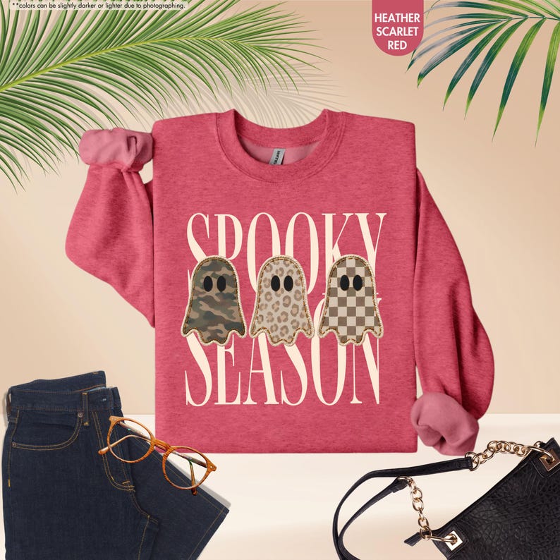 May include: A heather scarlet red crewneck sweatshirt featuring the white text SPOOKY SEASON and three ghost illustrations with camouflage, leopard print, and checkerboard patterns. The garment is displayed with dark blue jeans and a black bag.