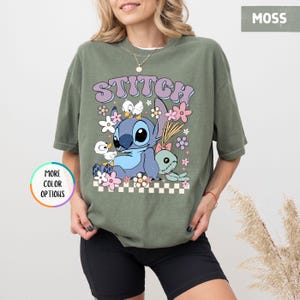 May include: Moss green t-shirt featuring a graphic of Stitch with the word "STITCH" above. The design includes Stitch, flowers, and other characters. The shirt has a relaxed fit and is made of a soft material.