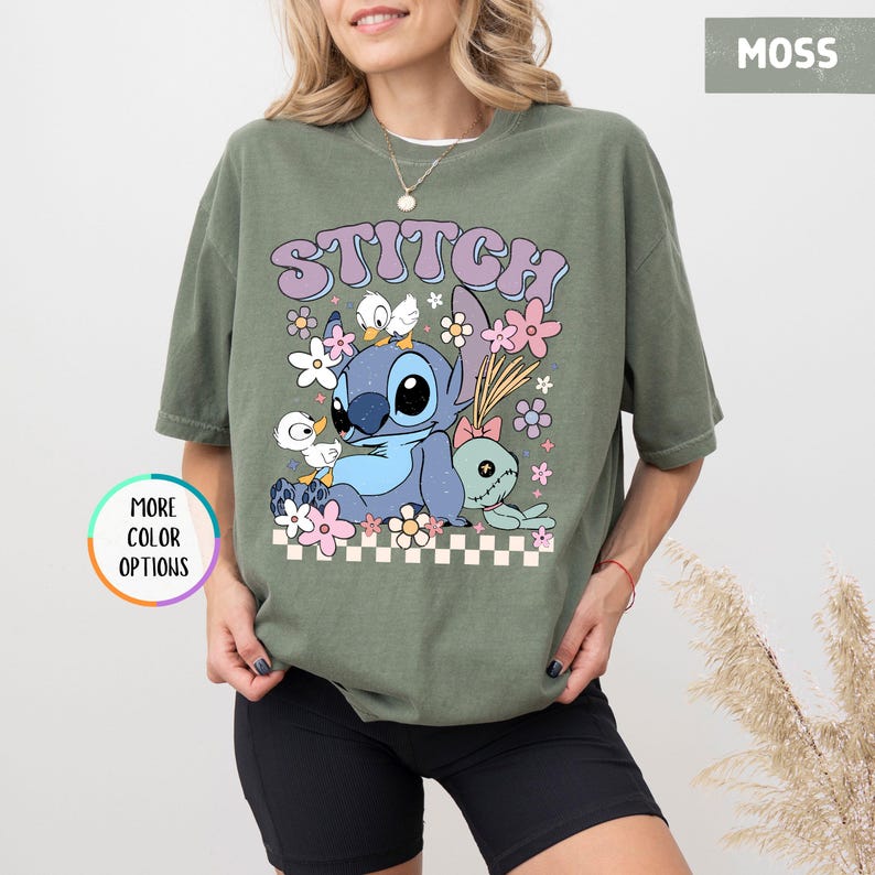 May include: Moss green t-shirt featuring a graphic of Stitch with the word "STITCH" above. The design includes Stitch, flowers, and other characters. The shirt has a relaxed fit and is made of a soft material.