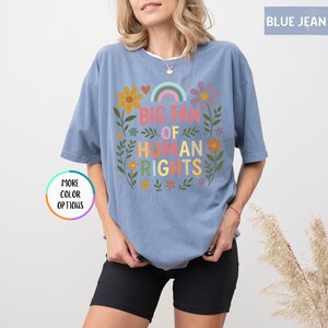 May include: Blue jean-colored t-shirt with the text "BIG FAN OF HUMAN RIGHTS" in colorful letters. The design includes a rainbow, flowers, and leaves. The shirt has short sleeves and a relaxed fit.