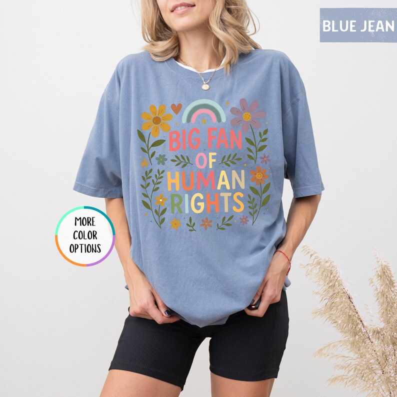 May include: Blue jean-colored t-shirt with the text "BIG FAN OF HUMAN RIGHTS" in colorful letters. The design includes a rainbow, flowers, and leaves. The shirt has short sleeves and a relaxed fit.