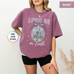 May include: Berry-colored t-shirt with the text "Happiest Mom on Earth" and a graphic of a castle, Mickey Mouse balloons, and a bow. The shirt is a relaxed fit and has short sleeves.