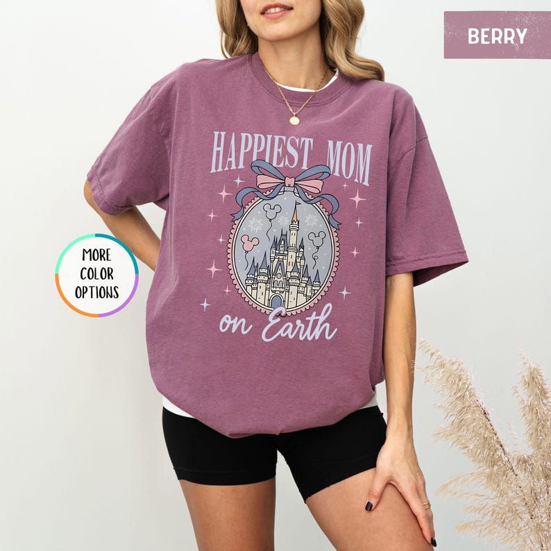 May include: Berry-colored t-shirt with the text "Happiest Mom on Earth" and a graphic of a castle, Mickey Mouse balloons, and a bow. The shirt is a relaxed fit and has short sleeves.