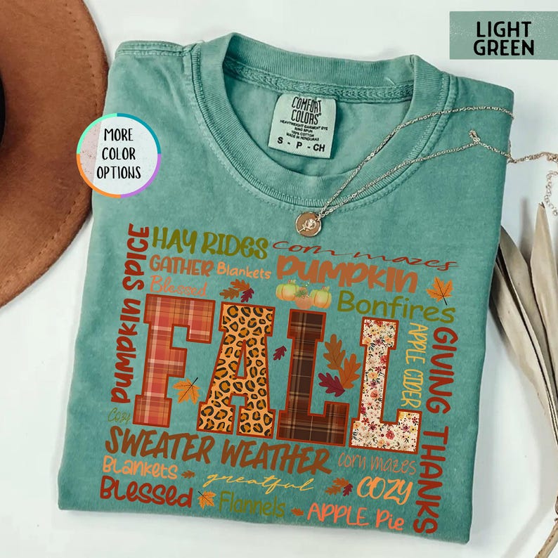 以下が含まれることがあります： 秋をテーマにしたデザインの、ライトグリーンのComfort Colors Tシャツ。「FALL」の文字が様々なパターンで大きく表示され、「Pumpkin Spice」、「Sweater Weather」、「Giving Thanks」などの言葉が周りに配置されています。シルバーのネックレスがTシャツの上にかけられています。