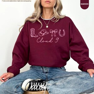 May include: Maroon sweatshirt featuring a light pink graphic of a cowboy boot, hat, bow, and horseshoe, with the text "LOTU Cloud 9". The sweatshirt is a solid maroon colour.