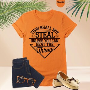 May include: Orange t-shirt with the text "Thou Shall Not Steal Unless You Can Beat The Throw" in black. The shirt is paired with blue jeans, eyeglasses, and cream-colored loafers.