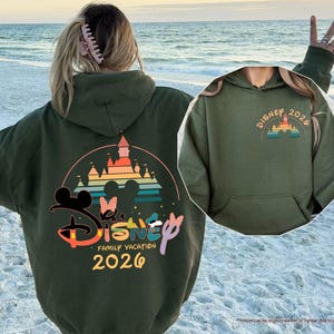 May include: An olive green hooded sweatshirt with a large graphic on the back featuring the Disney logo, Mickey and Minnie Mouse silhouettes, a castle, and the text "Disney Family Vacation 2026". A smaller graphic is on the front.