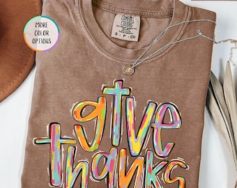 May include: Espresso-colored t-shirt with the words "give thanks" in a colorful, outlined font. The shirt has a crew neck and short sleeves. A silver necklace with a small pendant is draped across the shirt. The shirt is a Comfort Colors brand.