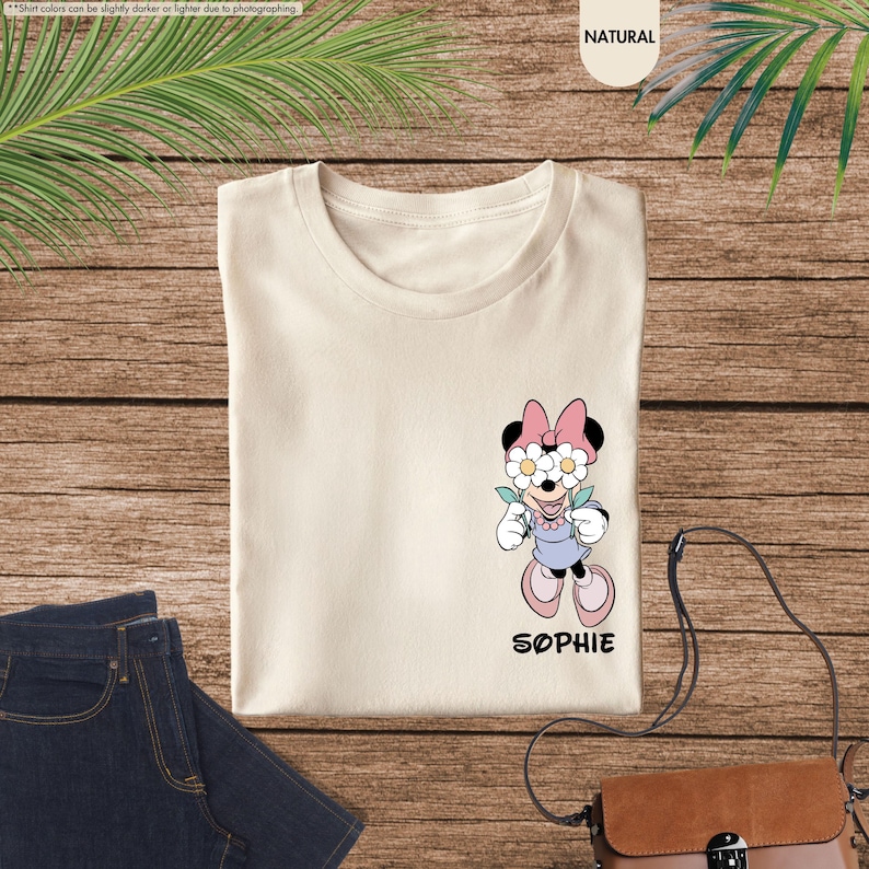 May include: Cream-colored t-shirt featuring a Minnie Mouse graphic with daisy-covered eyes, a pink bow, and a lavender dress. The name "SOPHIE" is printed below the character. The shirt is displayed on a wooden surface with a pair of blue jeans.