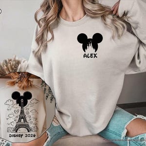 May include: A sand-colored crewneck sweatshirt featuring a black graphic on the left chest of a Mickey Mouse silhouette containing a castle and the name ALEX. The back displays a black graphic of the Eiffel Tower inside a Mickey silhouette.