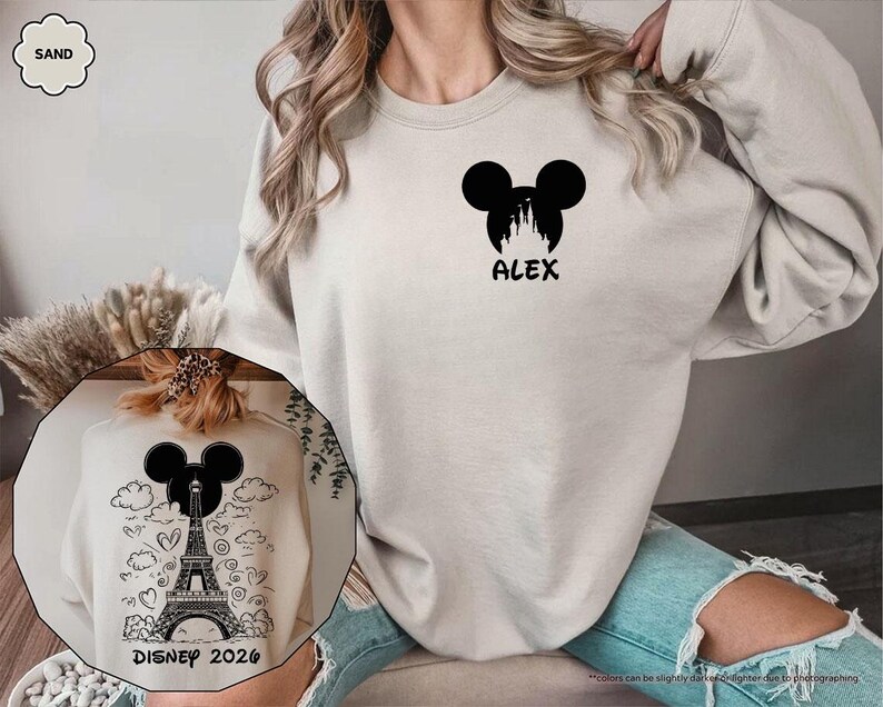 May include: A sand-colored crewneck sweatshirt featuring a black graphic on the left chest of a Mickey Mouse silhouette containing a castle and the name ALEX. The back displays a black graphic of the Eiffel Tower inside a Mickey silhouette.