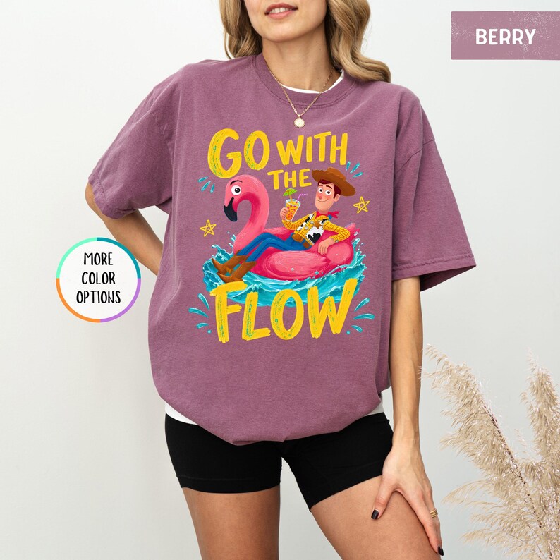 Puede incluir: Una camiseta de manga corta color baya con un gr&aacute;fico de un vaquero descansando sobre un flotador de flamenco rosa. El texto "GO WITH THE FLOW" aparece en letras amarillas. Una insignia circular a la izquierda dice "MORE COLOR OPTIONS".