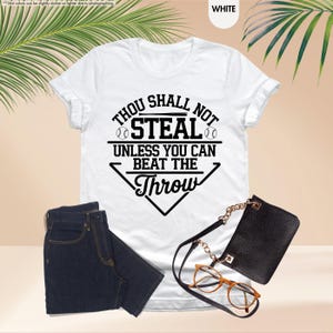May include: White t-shirt with the text "Thou Shall Not Steal Unless You Can Beat The Throw" in black. The shirt is paired with dark blue jeans, a black purse, and eyeglasses.
