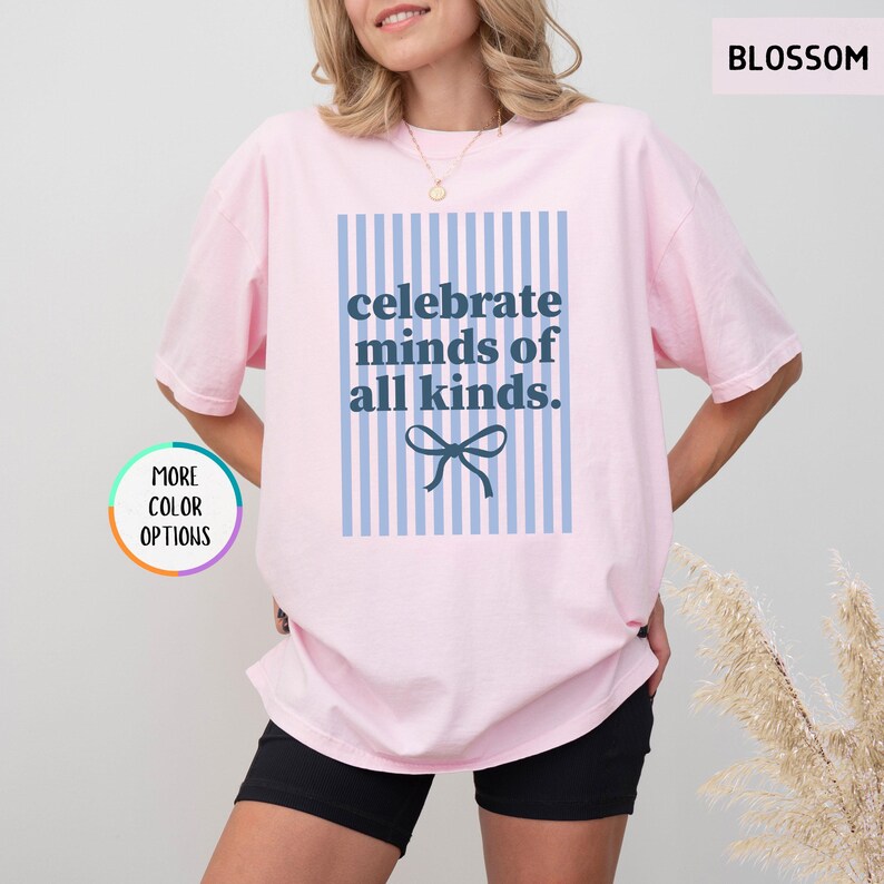 Puede incluir: Camiseta rosa claro con el texto azul "celebrate minds of all kinds" sobre un fondo de rayas azules y blancas. Un lazo azul est&aacute; debajo del texto. La camiseta es de corte holgado.