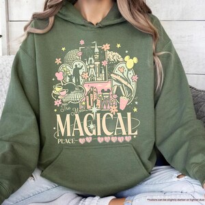 May include: Military green hoodie with a colorful graphic featuring a castle, Mickey Mouse, and the text "The Most Magical Place". The design includes pink, yellow, and white elements.