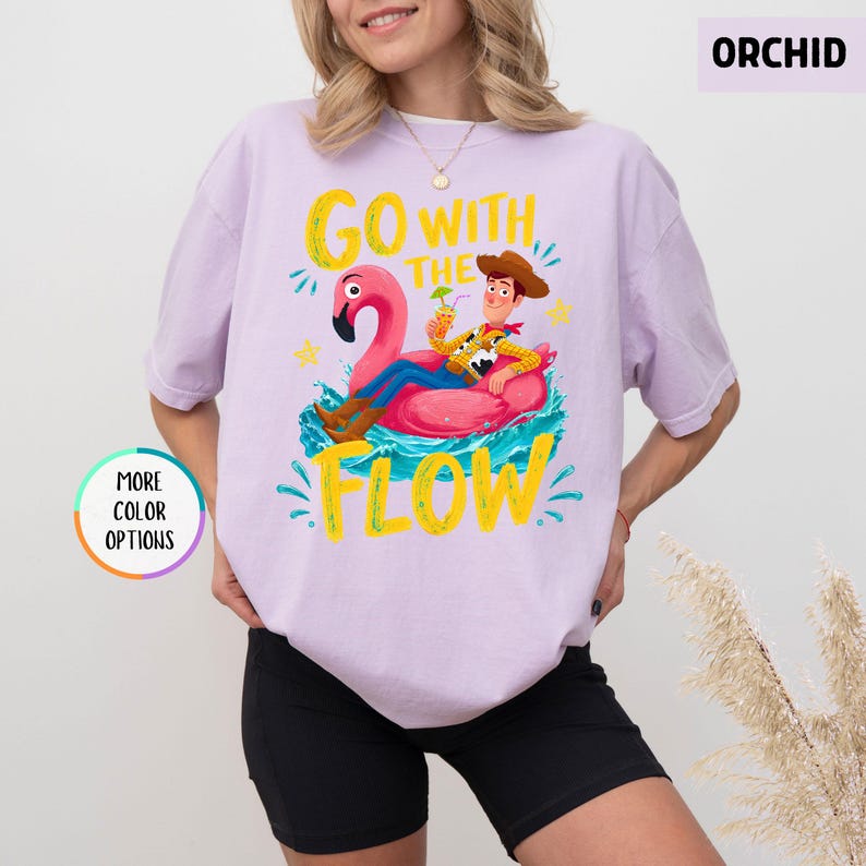 Puede incluir: Una camiseta color orqu&iacute;dea con un gr&aacute;fico de un vaquero de dibujos animados montado en un flotador de flamenco rosa en agua azul. El texto dice GO WITH THE FLOW en letras amarillas. El dise&ntilde;o incluye una bebida, salpicaduras y estrellas.