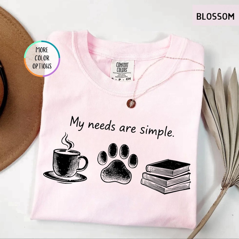 May include: A light pink t-shirt with the phrase "My needs are simple." and illustrations of a coffee cup, paw print, and books. The shirt has a crew neck and a Comfort Colors tag.
