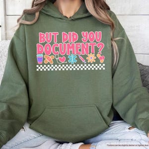 Puede incluir: Sudadera con capucha verde militar con el texto "BUT DID YOU DOCUMENT?" en rosa y blanco. El dise&ntilde;o incluye iconos m&eacute;dicos como una tirita, un coraz&oacute;n y una jeringa. Un patr&oacute;n de cuadros blancos y negros en la parte inferior.