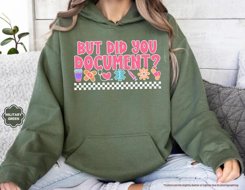 Puede incluir: Sudadera con capucha verde militar con el texto "BUT DID YOU DOCUMENT?" en rosa y blanco. El dise&ntilde;o incluye iconos m&eacute;dicos como una tirita, un coraz&oacute;n y una jeringa. Un patr&oacute;n de cuadros blancos y negros en la parte inferior.