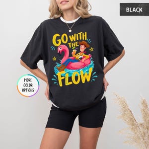 Puede incluir: Una camiseta negra con un gr&aacute;fico de un vaquero sentado en un flotador de flamenco rosa sobre agua azul. El texto "GO WITH THE FLOW" est&aacute; impreso en amarillo encima y debajo del gr&aacute;fico. Peque&ntilde;as estrellas amarillas rodean el dise&ntilde;o.