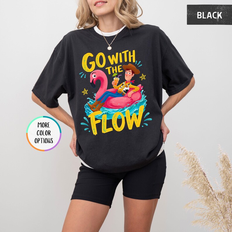 Puede incluir: Una camiseta negra con un gr&aacute;fico de un vaquero sentado en un flotador de flamenco rosa sobre agua azul. El texto "GO WITH THE FLOW" est&aacute; impreso en amarillo encima y debajo del gr&aacute;fico. Peque&ntilde;as estrellas amarillas rodean el dise&ntilde;o.