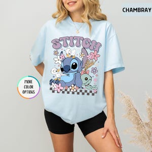 May include: Light blue t-shirt featuring a graphic of Stitch, ducks, and flowers. The word "STITCH" is printed above the graphic in a purple, retro font. The shirt has short sleeves and a crew neck.