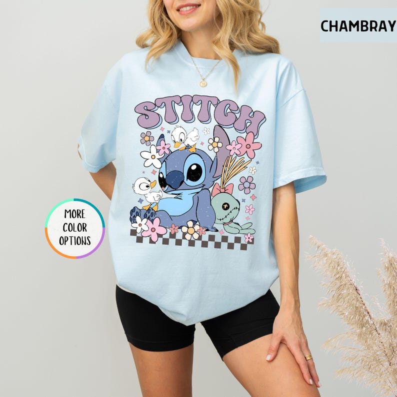 May include: Light blue t-shirt featuring a graphic of Stitch, ducks, and flowers. The word "STITCH" is printed above the graphic in a purple, retro font. The shirt has short sleeves and a crew neck.