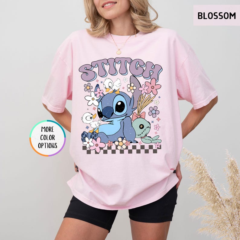 May include: A light pink t-shirt featuring a cartoon graphic of Stitch surrounded by flowers and the word "STITCH" in a retro font. The design includes a checkered pattern at the bottom. The shirt has a relaxed fit.