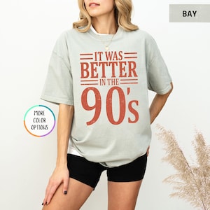 May include: A sage green short-sleeve t-shirt featuring the text IT WAS BETTER IN THE 90's printed in a distressed rust-orange font. The shirt has a crew neckline and a relaxed fit, paired with black shorts.