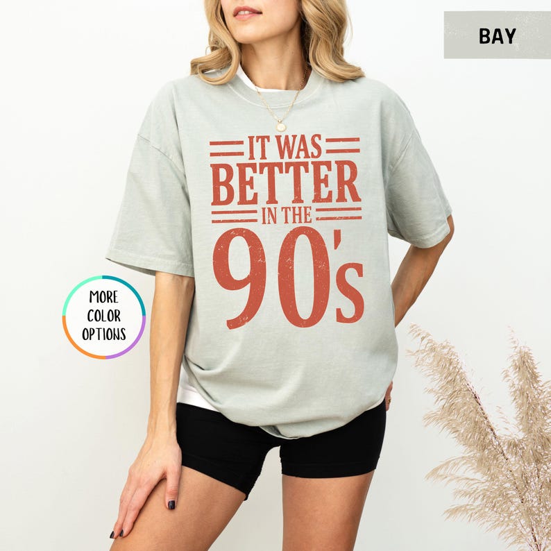 May include: A sage green short-sleeve t-shirt featuring the text IT WAS BETTER IN THE 90's printed in a distressed rust-orange font. The shirt has a crew neckline and a relaxed fit, paired with black shorts.