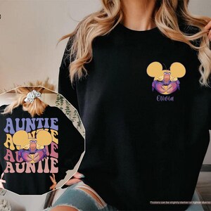 May include: Black sweatshirt featuring a cartoon crab design with yellow ears and the name "Olivia" in purple. The back of the sweatshirt has the word "AUNTIE" in a retro font. The sweatshirt is a comfortable, casual style.