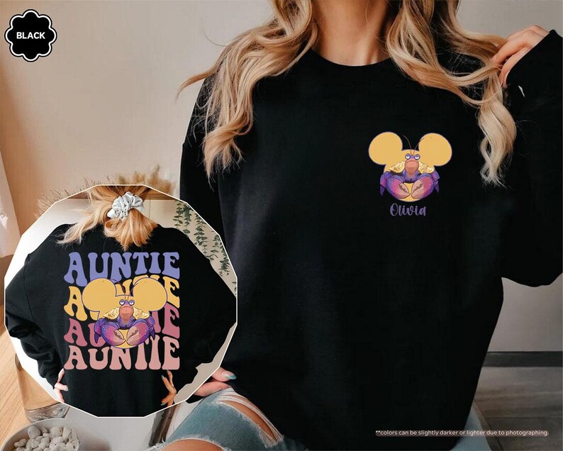 May include: Black sweatshirt featuring a cartoon crab design with yellow ears and the name "Olivia" in purple. The back of the sweatshirt has the word "AUNTIE" in a retro font. The sweatshirt is a comfortable, casual style.