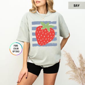 May include: A sage green short-sleeve t-shirt featuring a large red strawberry graphic with a green stem centered over horizontal blue stripes. The shirt has a crew neckline and a relaxed fit. A small circular graphic reads MORE COLOR OPTIONS.