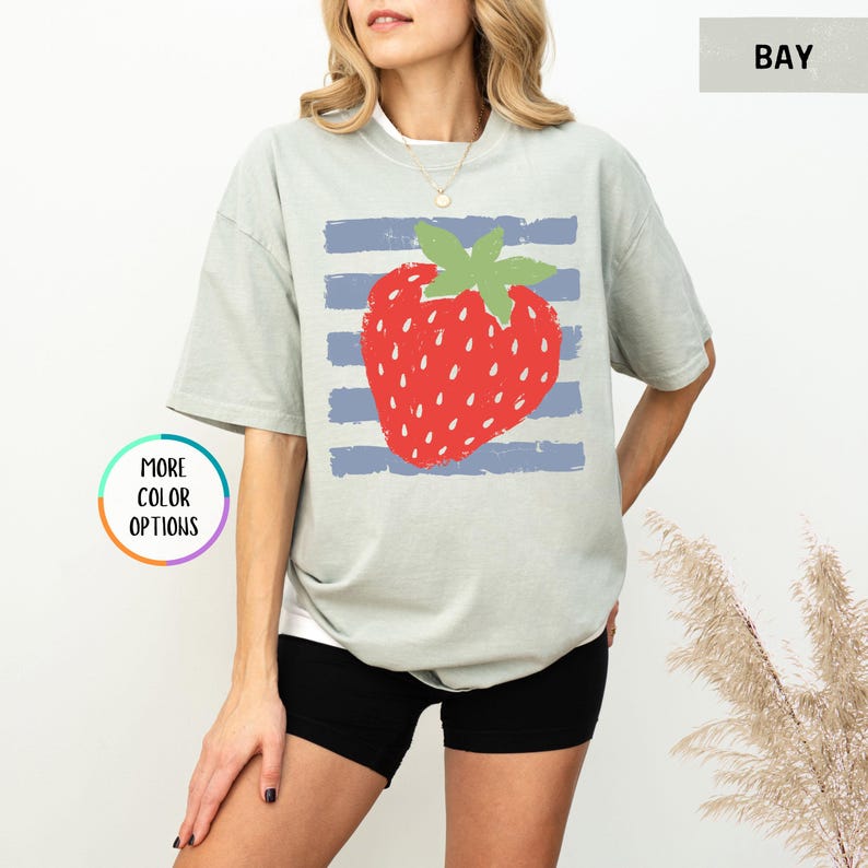 May include: A sage green short-sleeve t-shirt featuring a large red strawberry graphic with a green stem centered over horizontal blue stripes. The shirt has a crew neckline and a relaxed fit. A small circular graphic reads MORE COLOR OPTIONS.