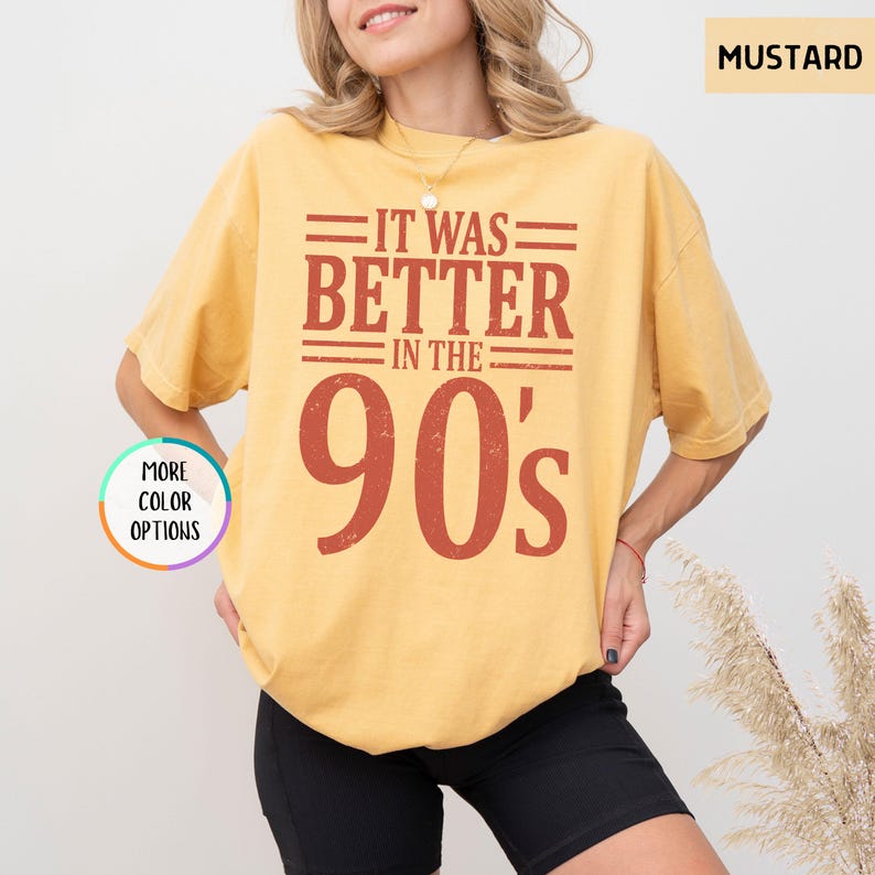 May include: A mustard yellow short-sleeve t-shirt featuring the dark red text IT WAS BETTER IN THE 90's centered on the chest. A small circular graphic on the left side contains the text MORE COLOR OPTIONS and the word MUSTARD appears in the top right.