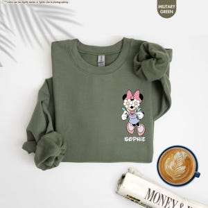 May include: Military green sweatshirt featuring a cartoon Minnie Mouse design with a floral headband and the name "Sophie" in white. The sweatshirt has long sleeves and a crew neck. A cup of coffee and a newspaper are in the background.